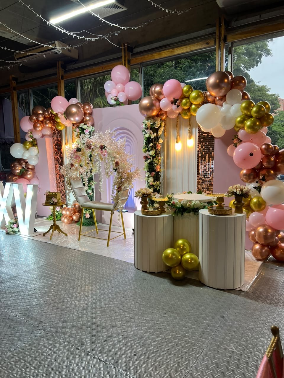 Montaje de quince años con arco de globos rosa, oro y blanco, flores y mesas